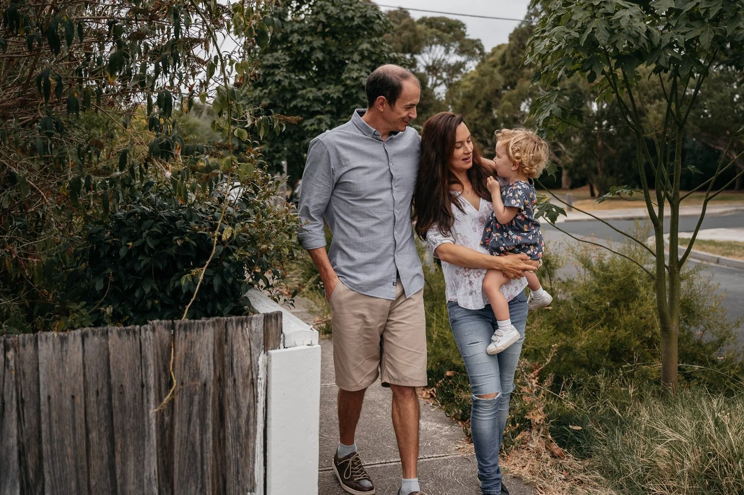 A family of three walking on a sidewalk in a suburban neighborhood, with the mother holding a young girl and the father walking beside them.
