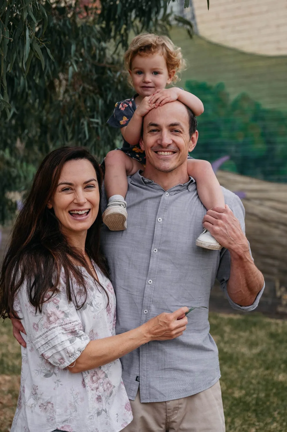 Smiling family of three outdoors, woman and man holding a young girl on the man's shoulders, trees and greenery in the background.