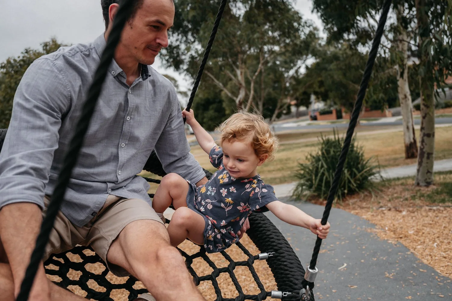 A man and a young girl sitting on a black rope swing outdoors in a park with trees and a walking path.