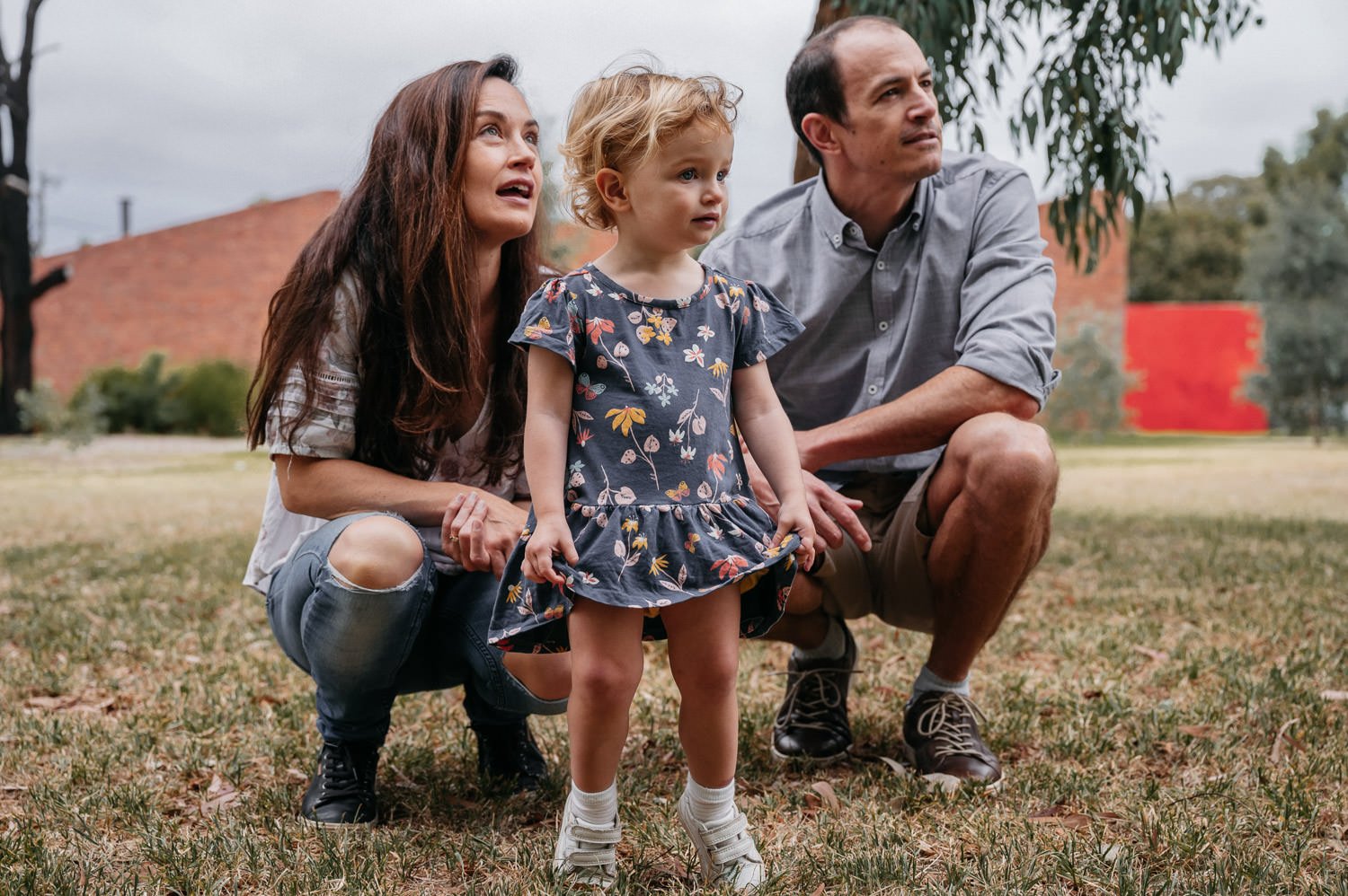 A family of three crouching on the grass, looking up with concerned expressions. The mother has long brown hair and is wearing a ripped jeans and a white top. The young girl has curly blonde hair and a floral dress. The father has short dark hair, we