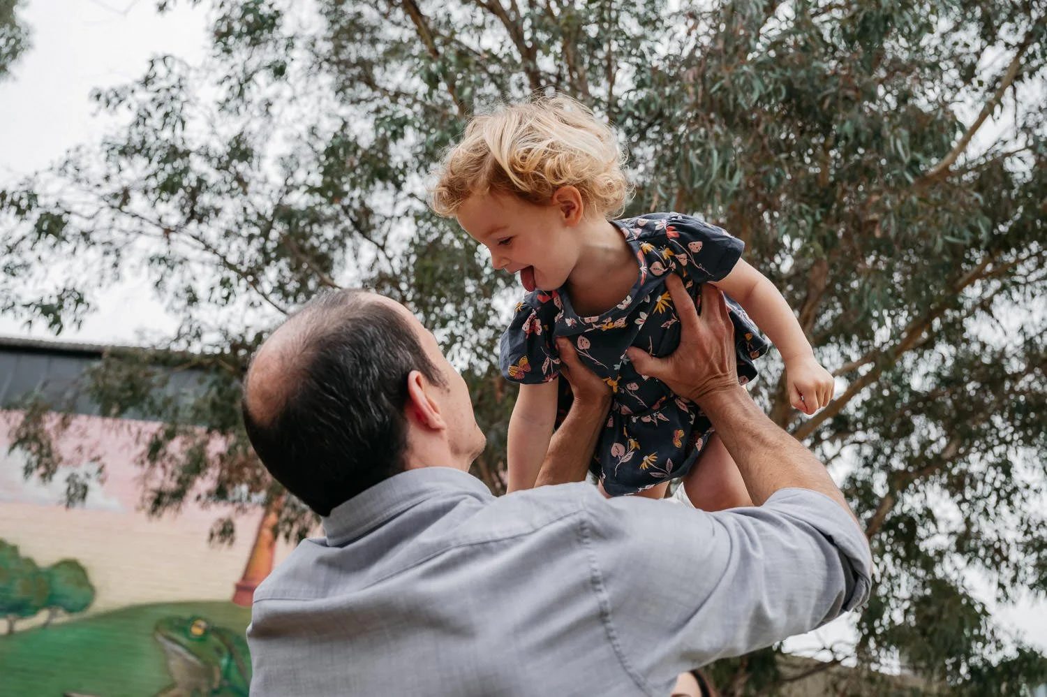 A man lifting a young girl, both smiling, outdoors with trees in the background.