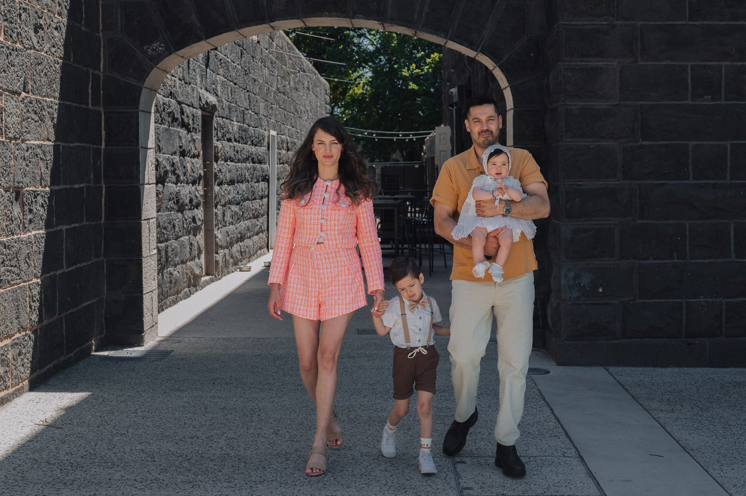 A family of four walking together outdoors, holding hands, under a stone archway. The mother is wearing a pink and orange checkered outfit, the father in a tan shirt and white pants holding a baby girl dressed in white, and their son in brown shorts 