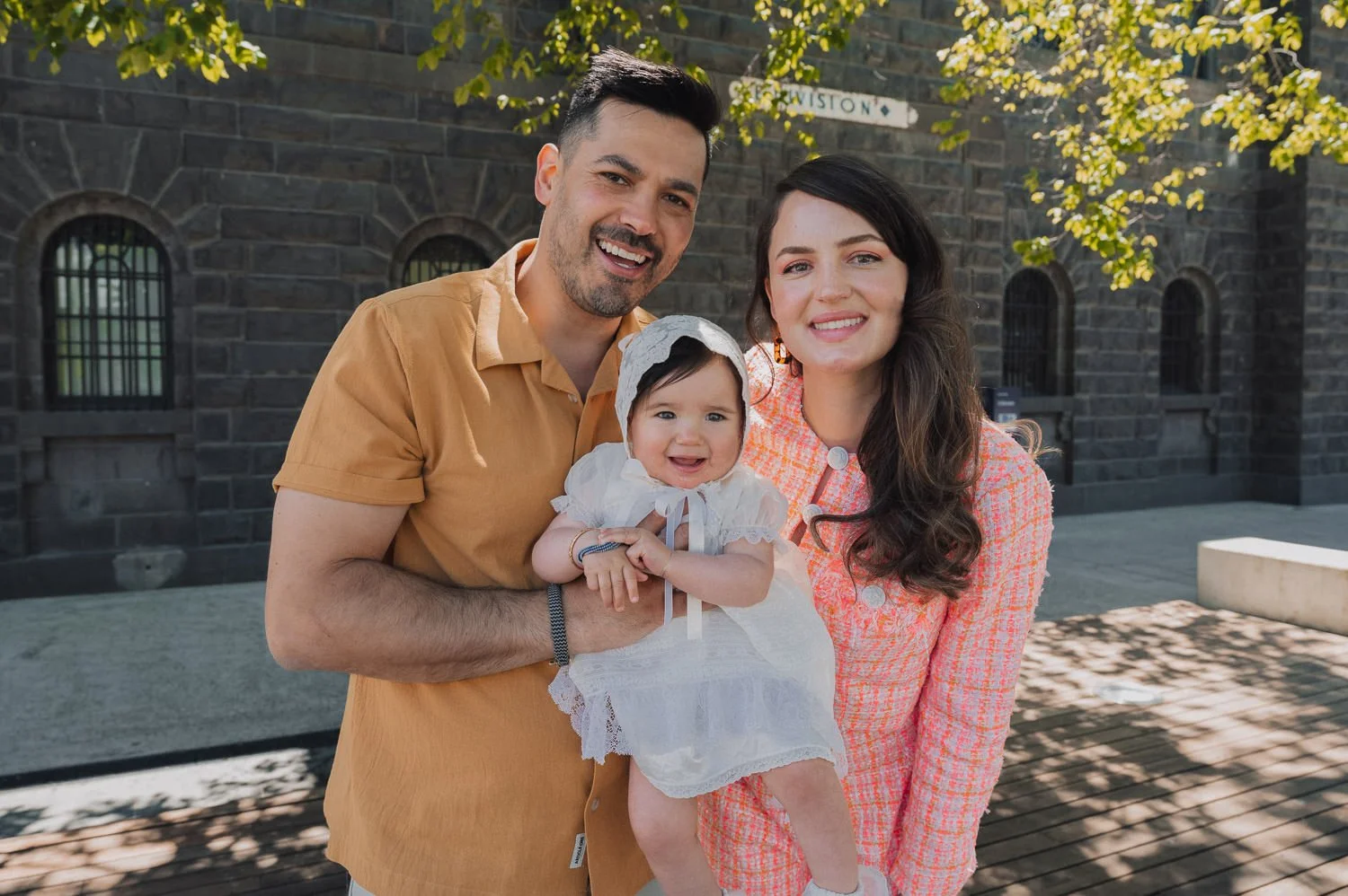 Young family of three smiling outdoors in front of a brick building, with a man, woman, and a baby girl dressed in white, all enjoying a sunny day.