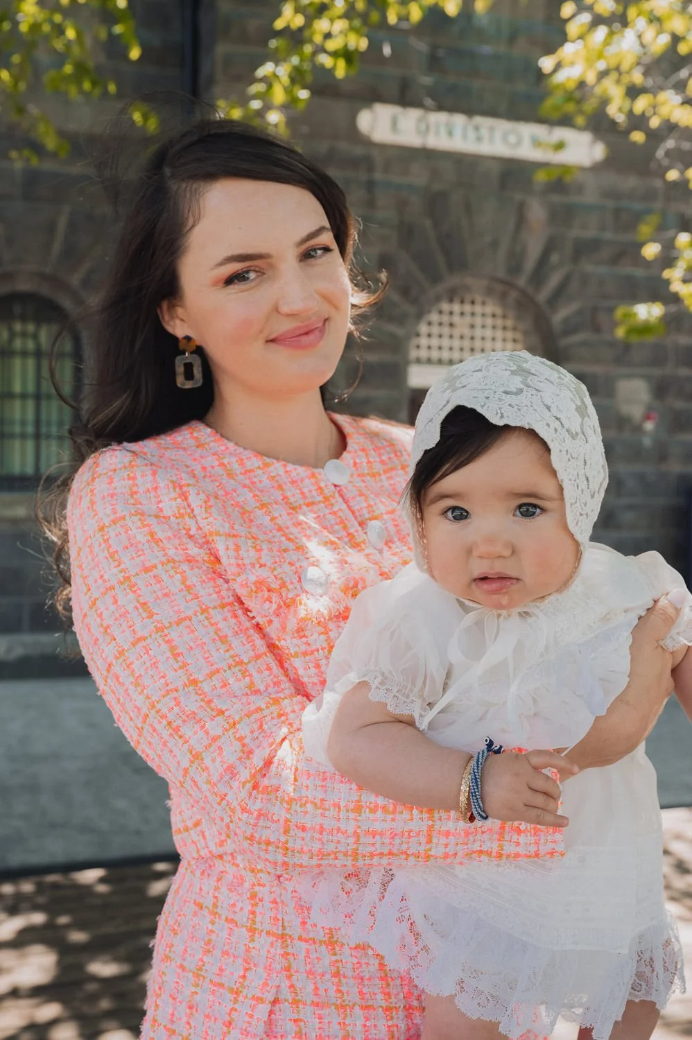 A woman holding a baby girl outdoors, in front of a brick building with trees and a sign. The woman is smiling, wearing a colorful dress and earrings, and the baby girl is dressed in white with a lace bonnet.