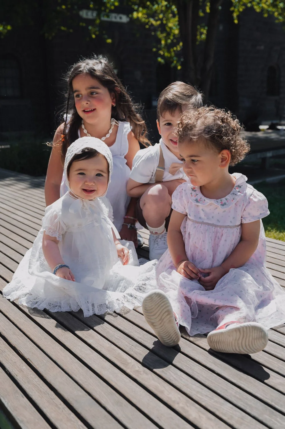 Five happy children outdoors on a wooden deck, dressed in light summer clothes, with a background of greenery and a stone building.