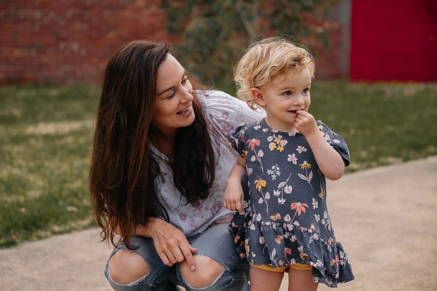 A woman with long dark hair and a child with curly blonde hair outdoors on a sidewalk, smiling and interacting with each other.