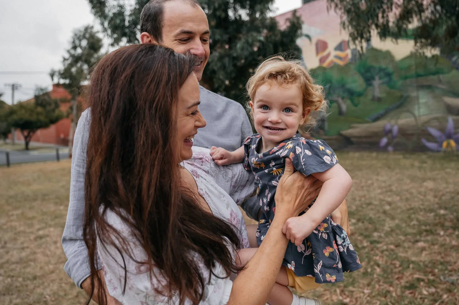 A woman and two men playing with a young girl outdoors in a park, with trees and mural artwork in the background.