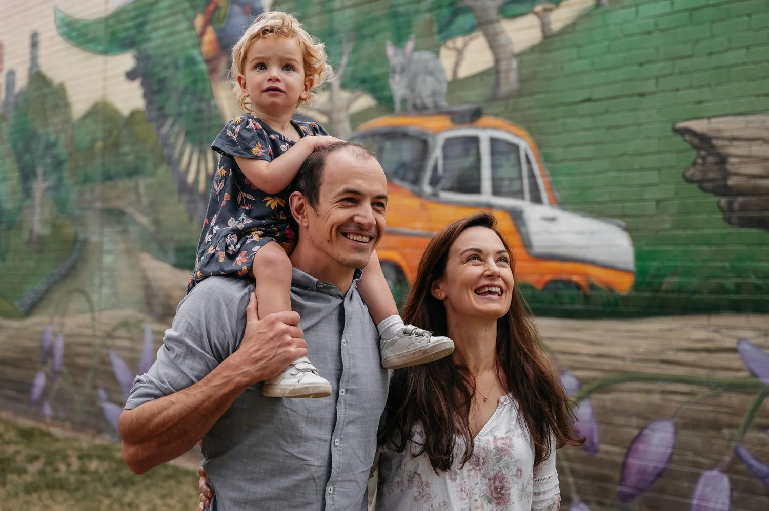 A happy family of three enjoying outdoors with a colorful mural of a truck and nature scene in the background. The father is carrying their young daughter on his shoulders, and the mother stands beside them smiling.
