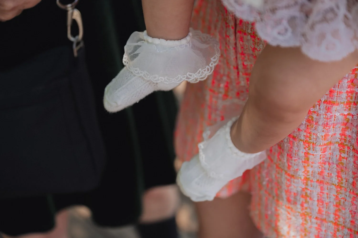 Close-up of a baby's legs wearing white lace socks and a pink dress with a colorful pattern, standing next to an adult wearing black pants.