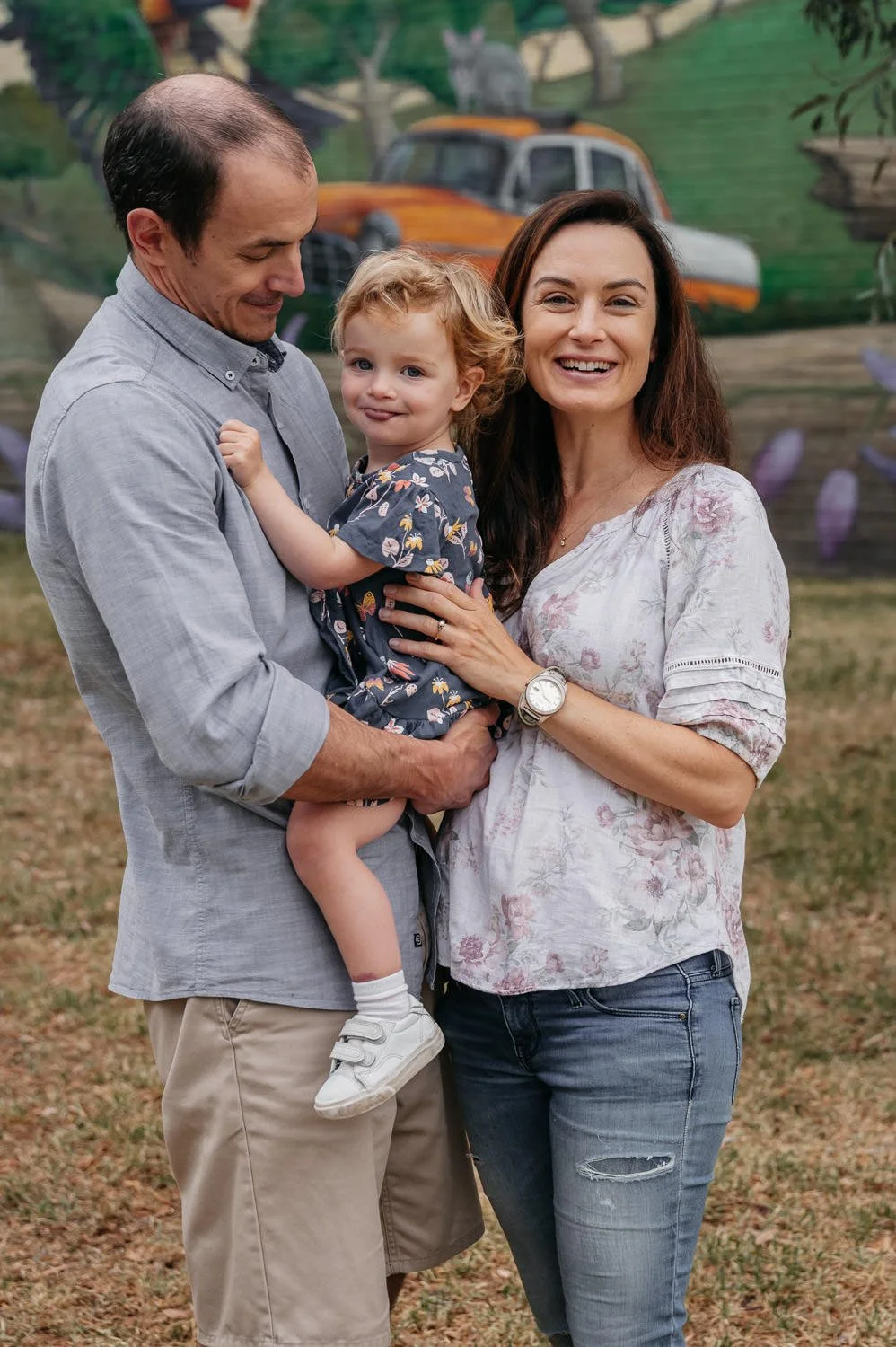 A family of three outdoors, with a man holding a young girl, and a woman standing beside them, in front of a colorful mural.