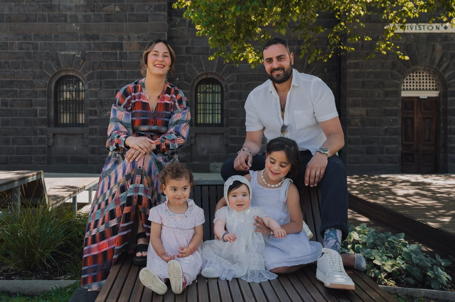 A family of six with two adults and four children, sitting outdoors in front of a dark brick building with arched windows and trees providing shade, smiling and posing for the photo.