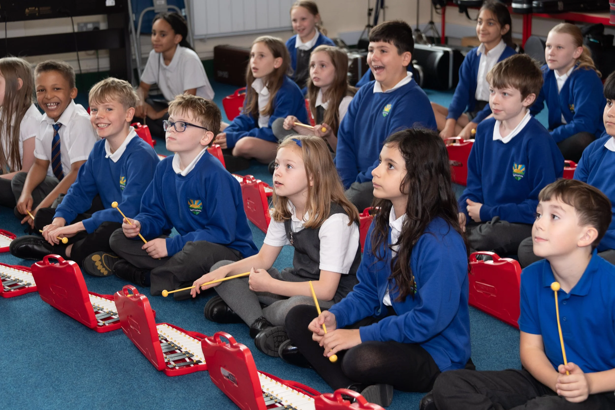 A group of school children, boys and girls, sitting cross-legged on a blue carpeted floor, playing xylophones in a classroom or music room.