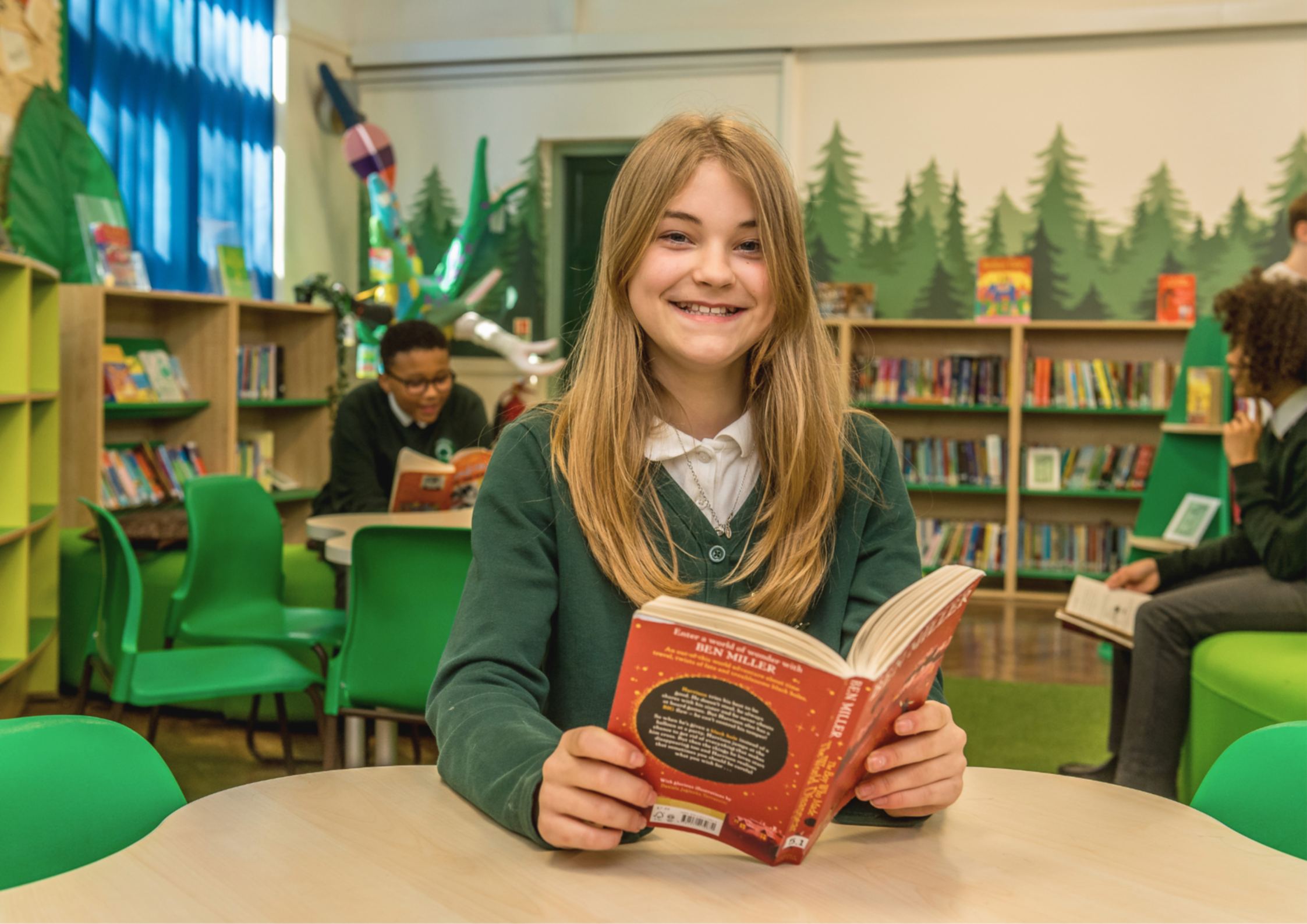 A young girl with long blonde hair is sitting at a table in a colorful library, smiling and reading a red book. Behind her, other children are reading or browsing books, with bookshelves and wall decorations, including trees and an imaginative dragon in the background.