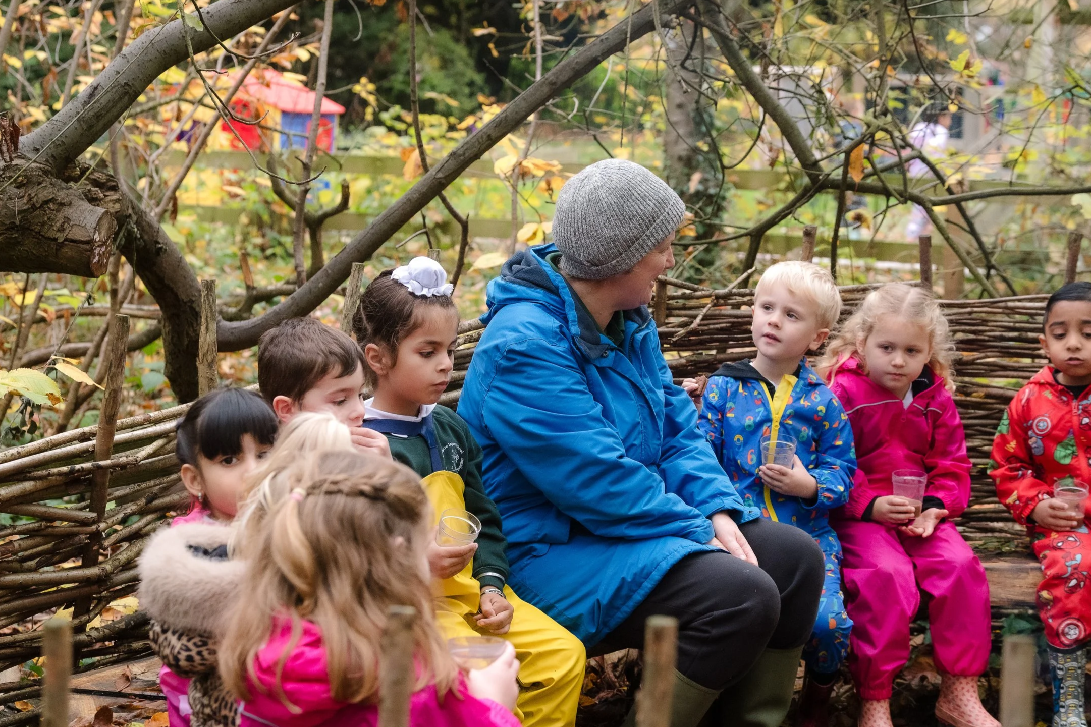 A group of children and an adult sitting outside on a wooden bench behind a woven wooden fence, surrounded by autumn leaves and trees, with some children holding drinks in cups.