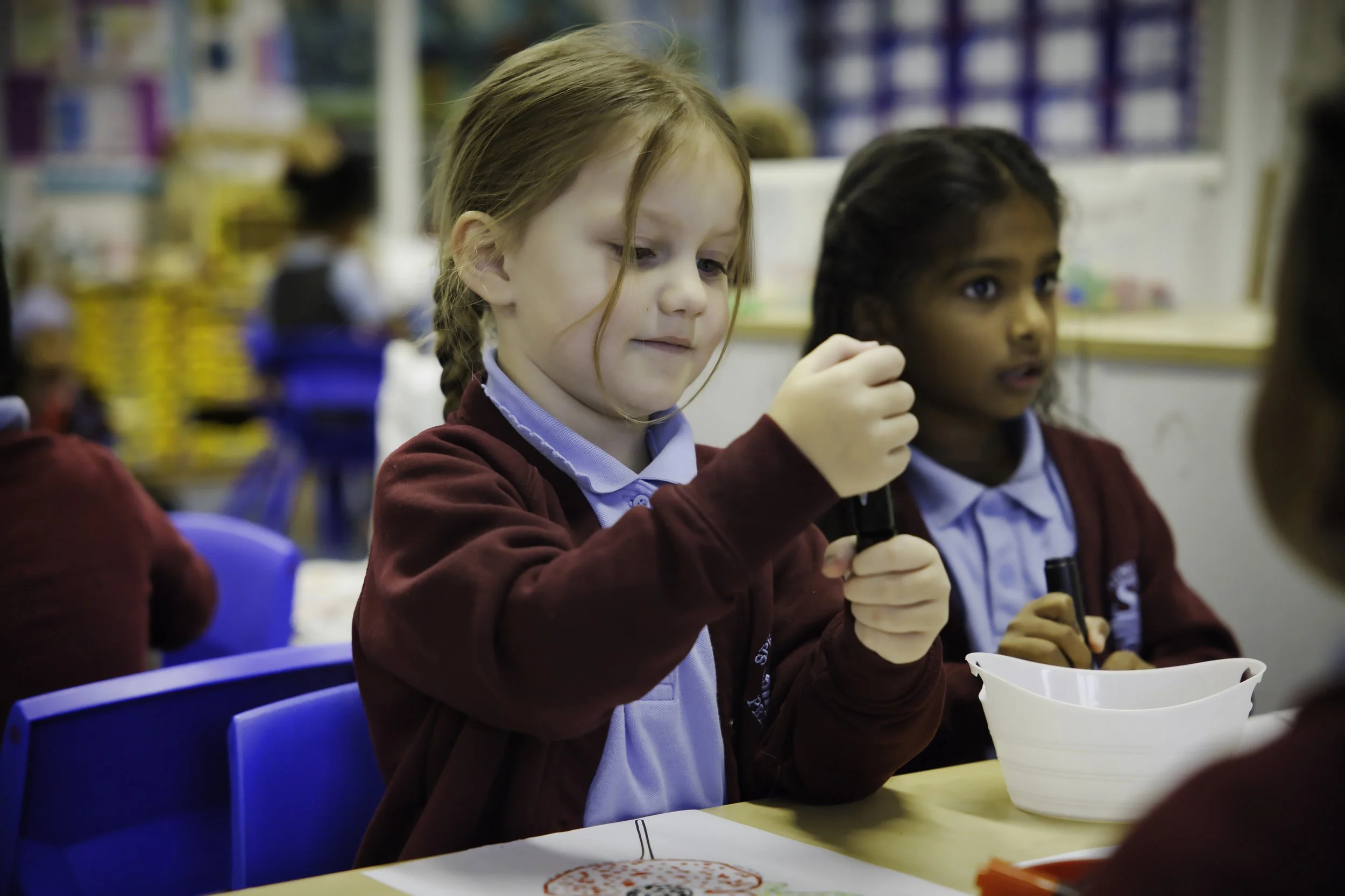 Young girl with red hair and a light blue school uniform in a classroom, focused on an activity with a white bowl on the table, other children and classroom materials in the background.