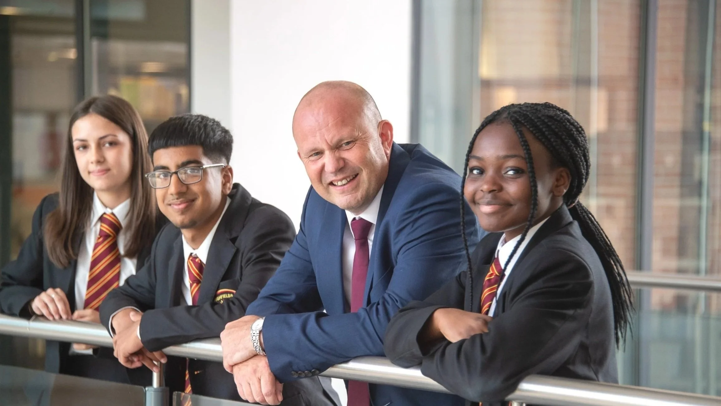 A diverse group of four students standing in a modern school or office environment, smiling and looking at the camera.