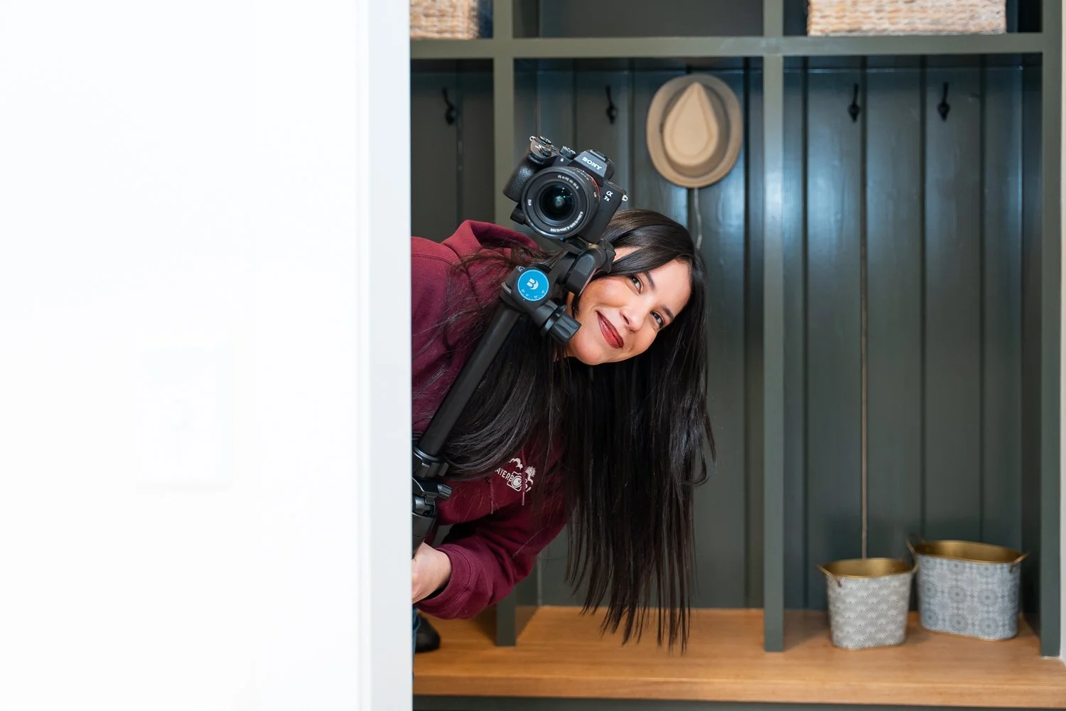 A woman with long dark hair and red lipstick peeking out from behind a white wall, holding a camera on a stabilizer, smiling at the camera. She is in a room with green shelving, two hats hanging on the wall, and two decorative baskets below.