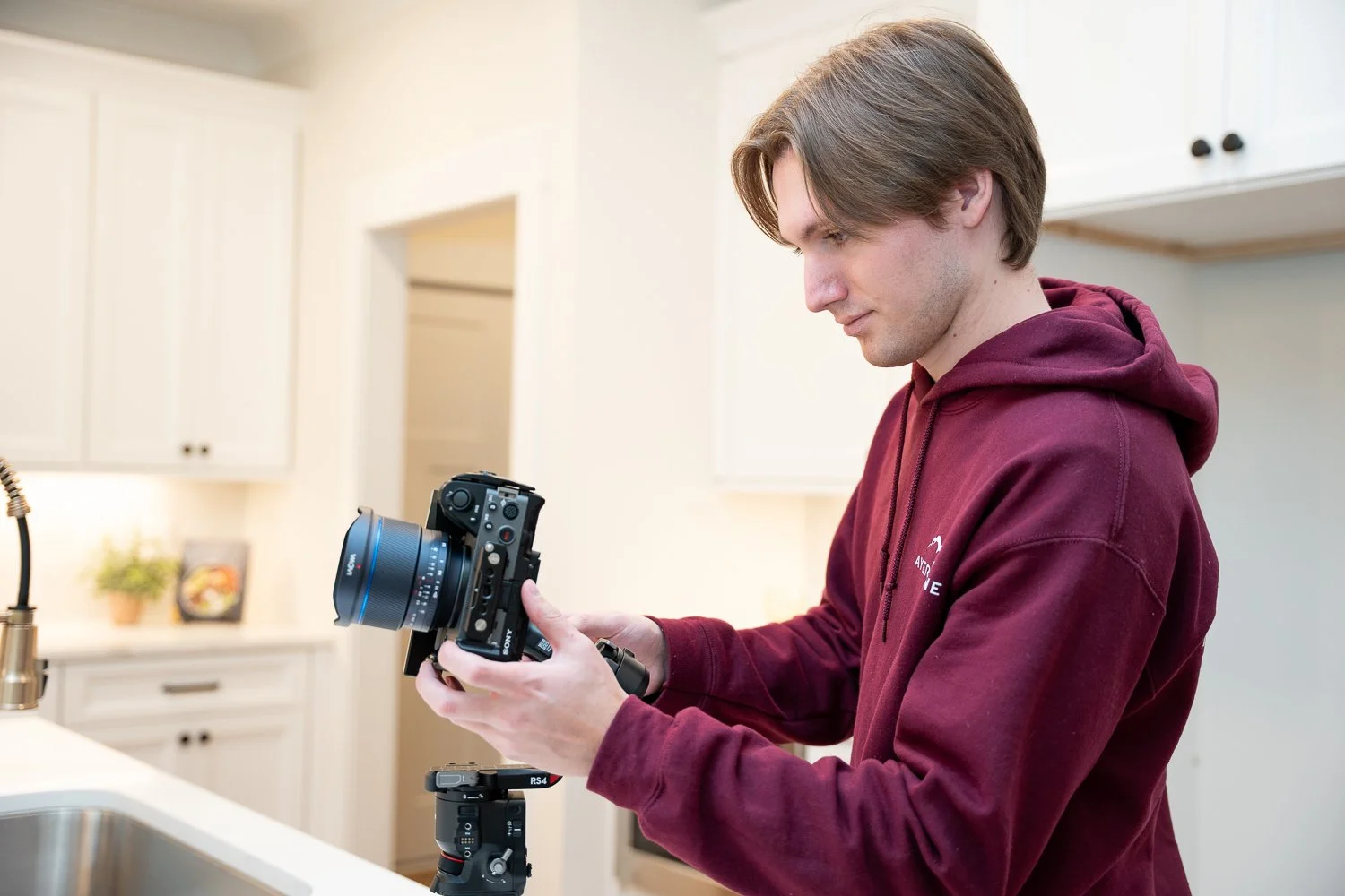 Young man with brown hair wearing a maroon hoodie holding a professional camera in a bright kitchen.