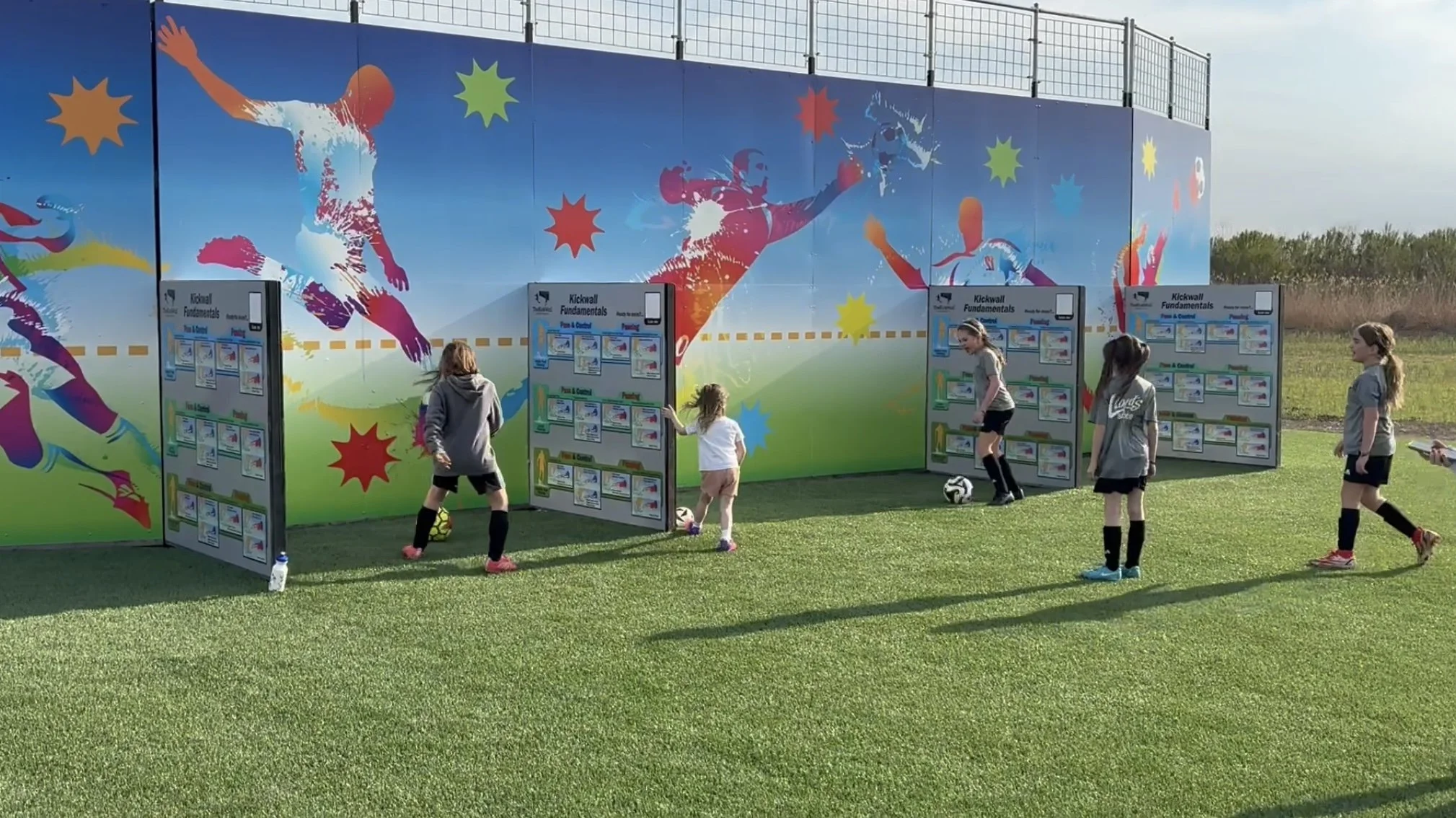 Children practicing soccer drills at a training station with colorful mural of soccer players on the wall.