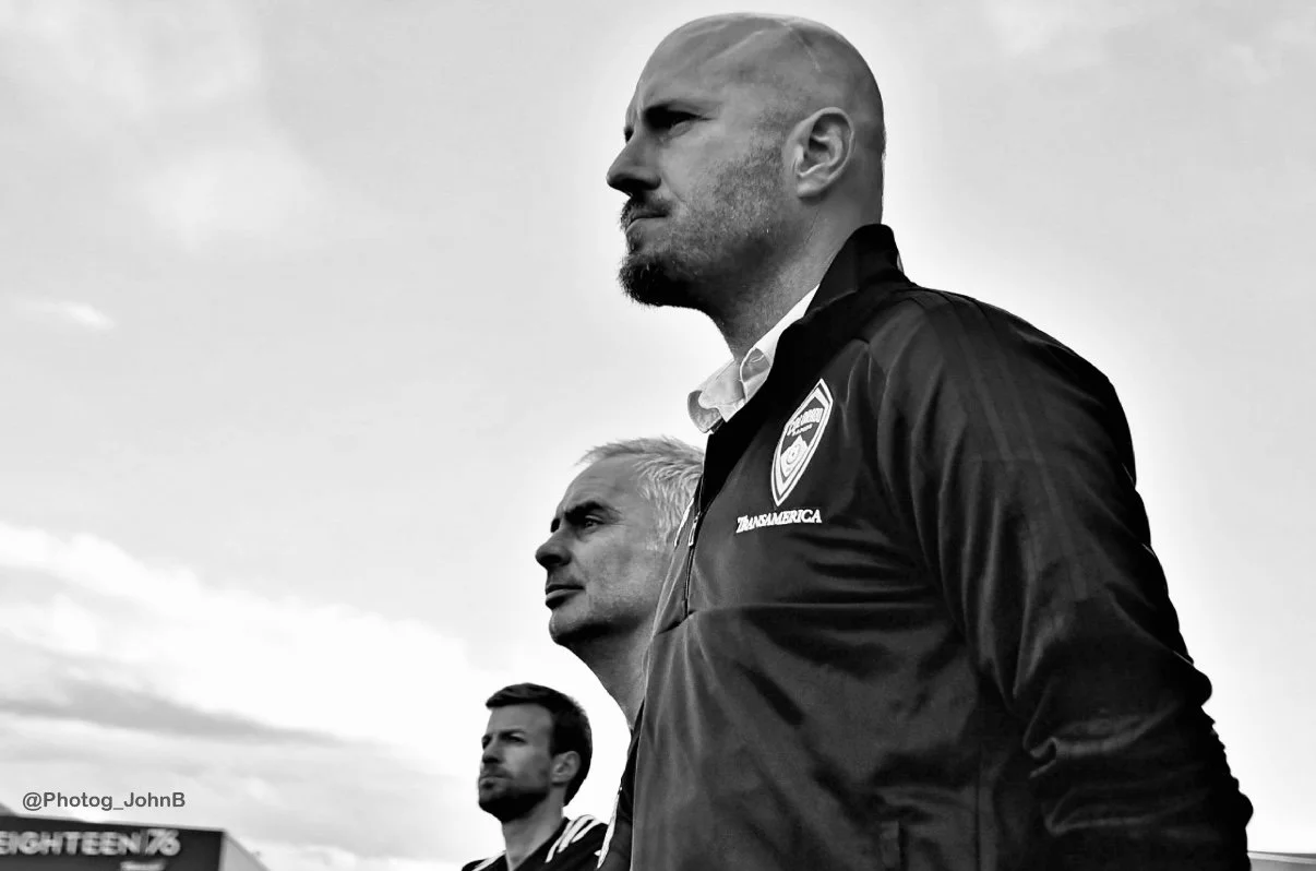 Three men standing outdoors looking into the distance, black and white photo, one man in the foreground wearing a jacket with a badge, other two in the background, cloudy sky.