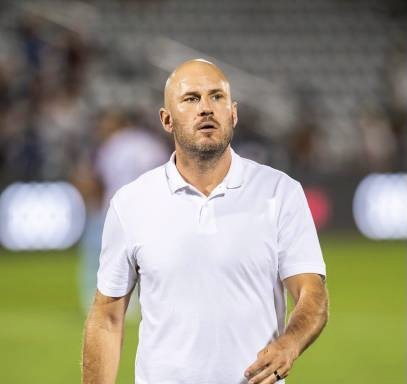 A bald man wearing a white polo shirt standing on a sports field with a stadium background.