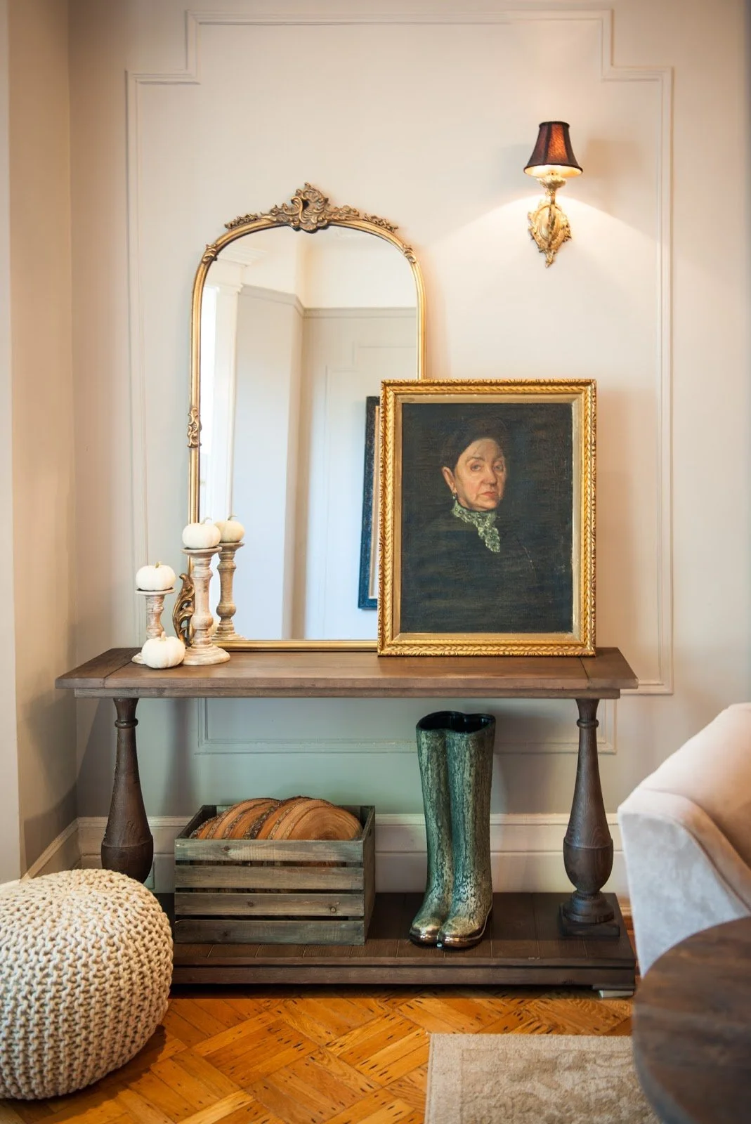 Interior decor with a wooden console table featuring a vintage portrait in a gold frame, a decorative mirror, white pumpkins on candlesticks, a rustic wooden crate, and a pair of tall boots. Beige walls and parquet flooring.