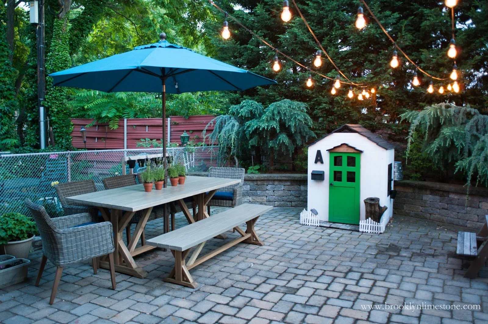 Cozy backyard patio with a wooden dining table, wicker chairs, and a blue umbrella. Potted plants decorate the table. String lights are hung overhead. A small playhouse with a green door is visible in the corner, surrounded by greenery.