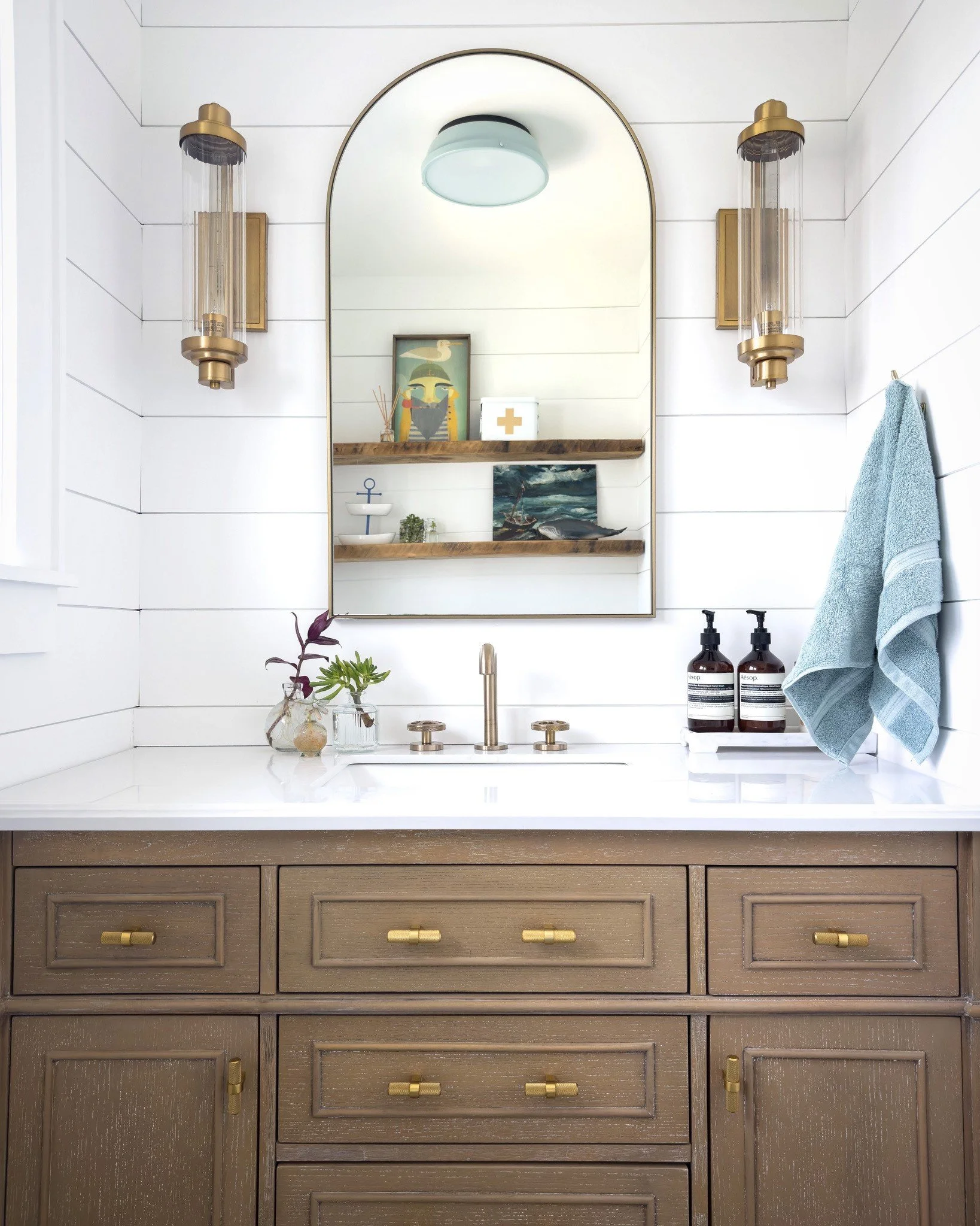 Bathroom vanity with wooden cabinet, white countertop, and brass fixtures, featuring an arched mirror flanked by modern light sconces, decorative shelves with artwork, and a hand towel.