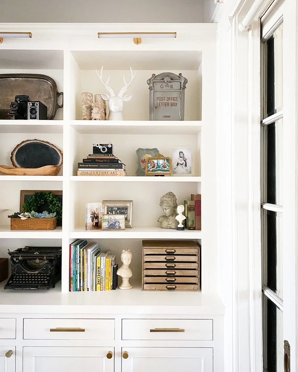 Decorative home bookshelf with vintage cameras, books, typewriter, letter box, and ornaments.
