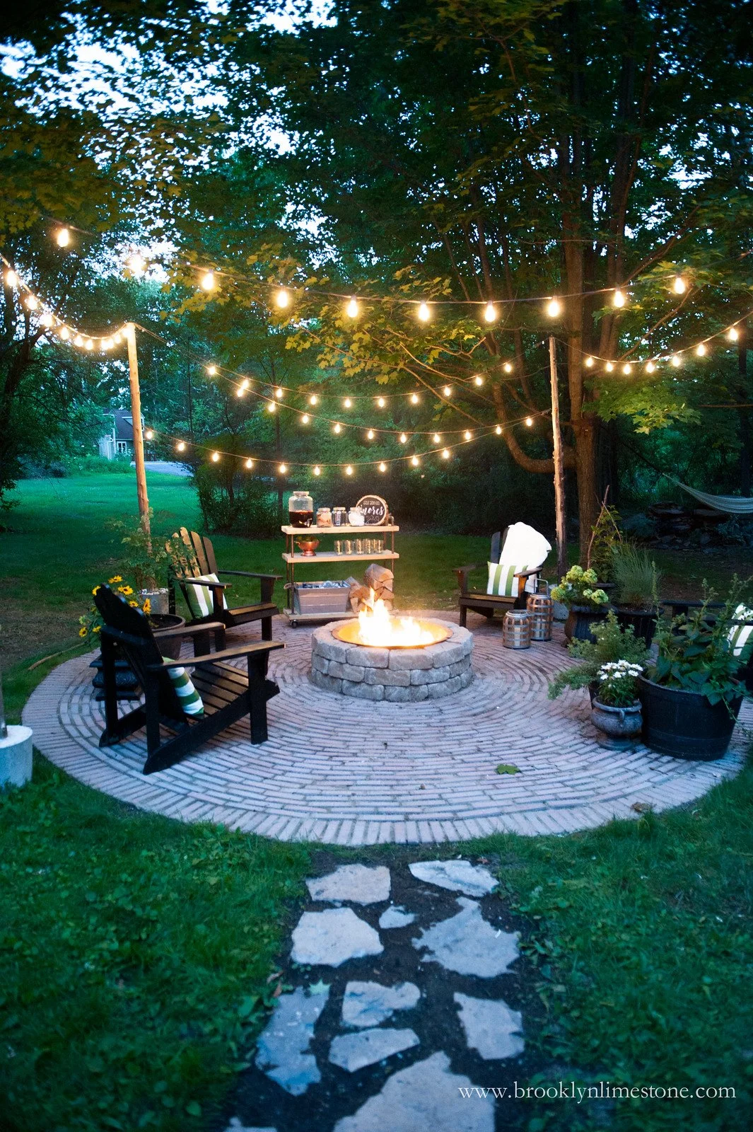 Outdoor patio with string lights and fire pit surrounded by chairs and plants at dusk.