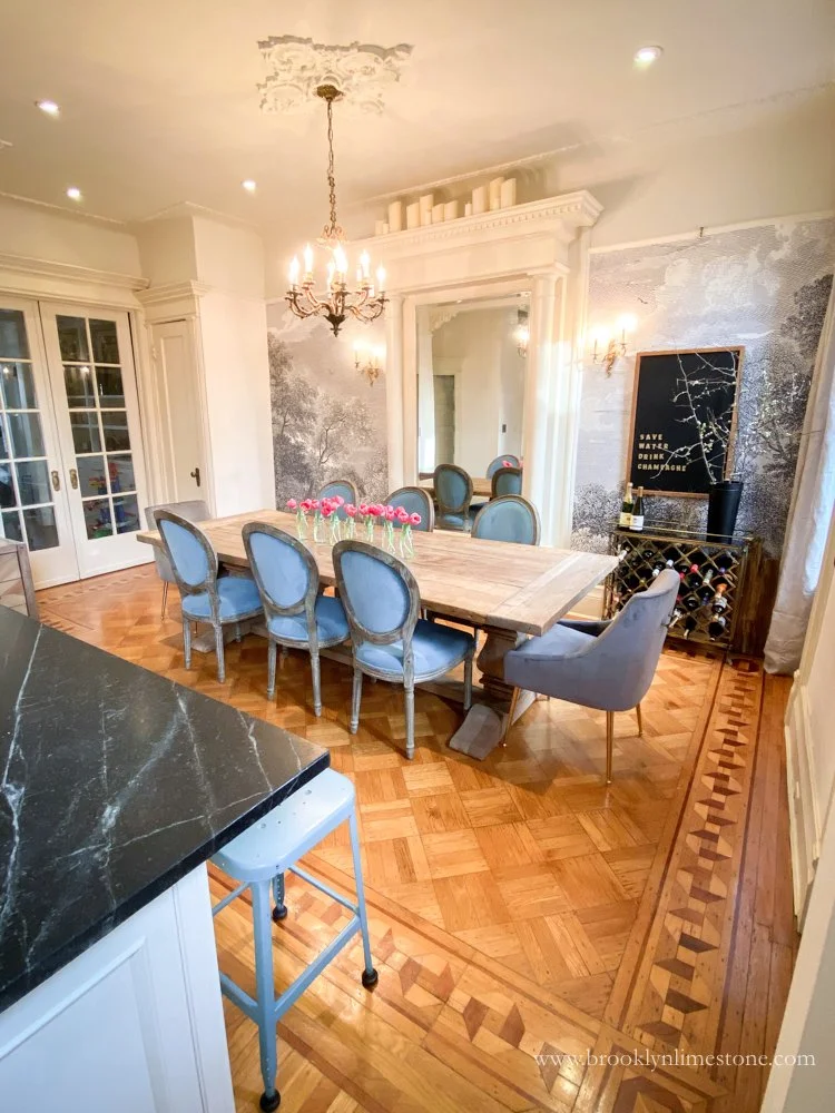 Elegant dining room with a large wooden table, blue chairs, chandelier, intricate wood flooring, and wall art. A kitchen counter with a stool is visible in the foreground.