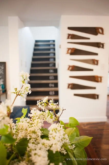 Interior view with staircase, vintage saws on wall, and white flowers in foreground.