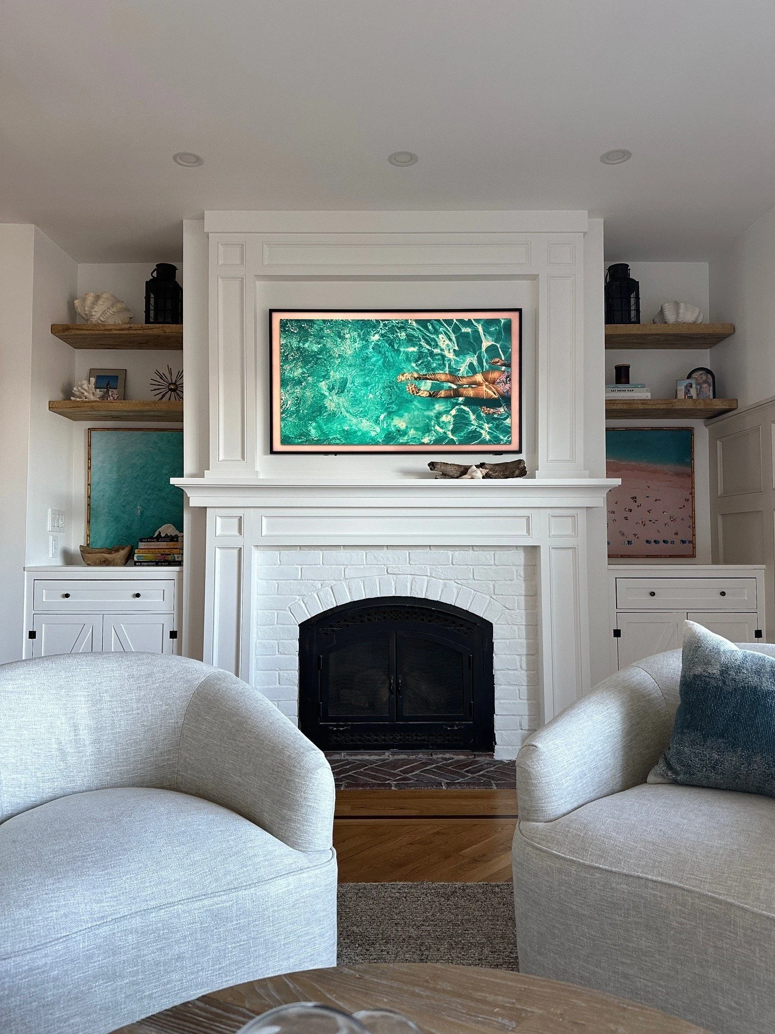 Living room with white fireplace, over which a TV displays a person swimming in a pool. Two beige armchairs and a round coffee table are in the foreground. White shelves with decorative items flank the fireplace.