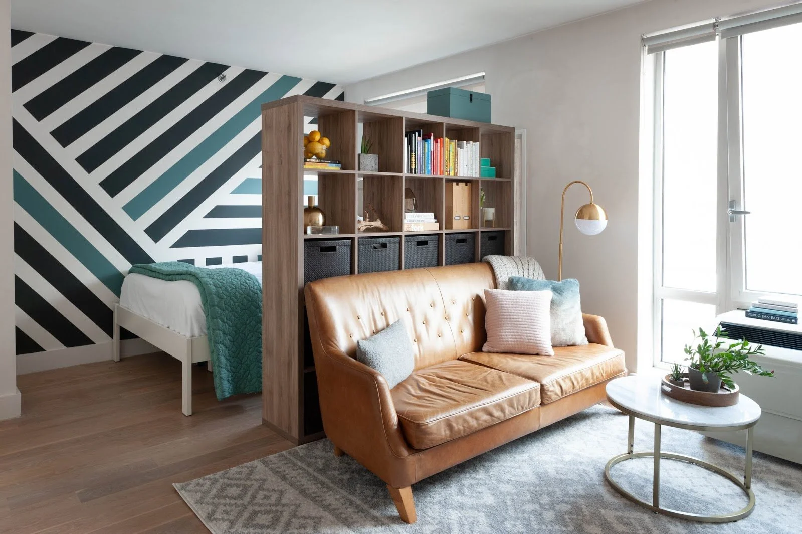 Modern studio apartment with leather sofa, geometric accent wall, wooden bookshelf, floor lamp, and a round coffee table with plants.