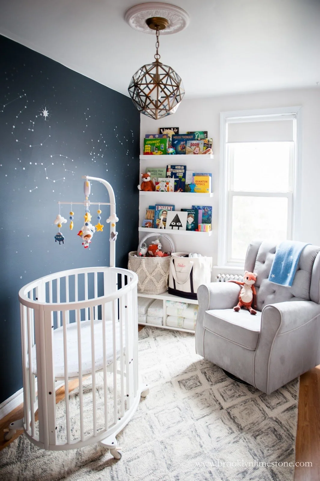 nursery room with white crib, mobile, grey armchair, bookshelf with children's books, dark wall with constellation mural, geometric pendant light, and toys