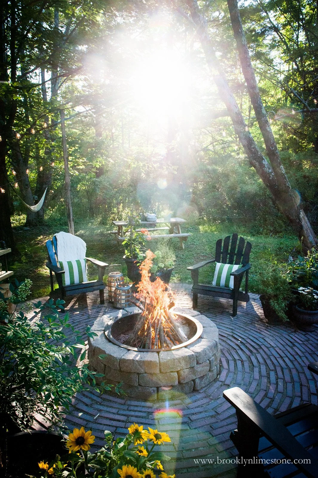 Outdoor patio with fire pit, surrounded by Adirondack chairs, plants, and sunflowers, with sunlight streaming through trees.