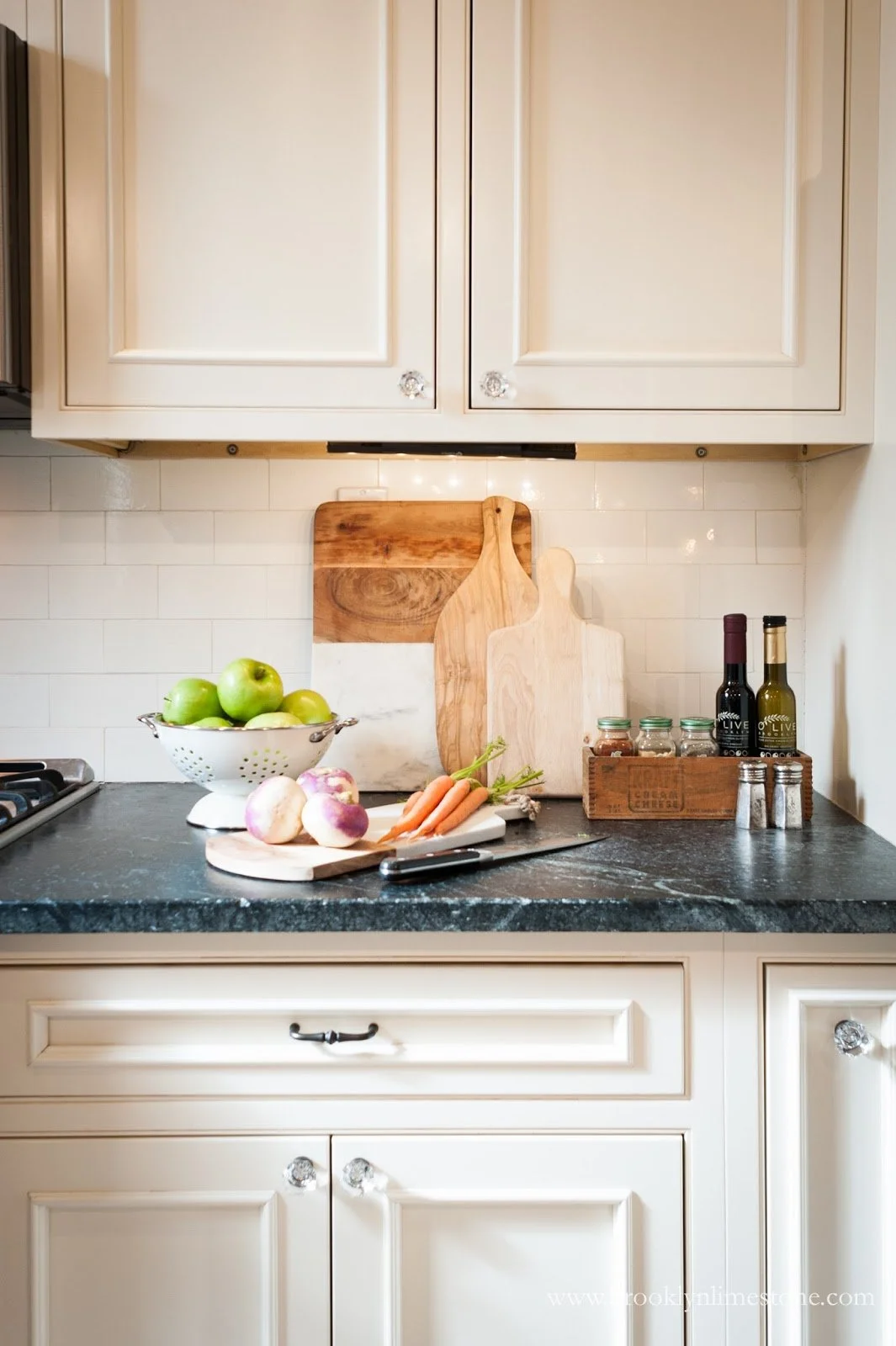 Kitchen countertop with cutting boards, green apples in a colander, carrots, turnips, olive oil bottles, spices, and white cabinets.