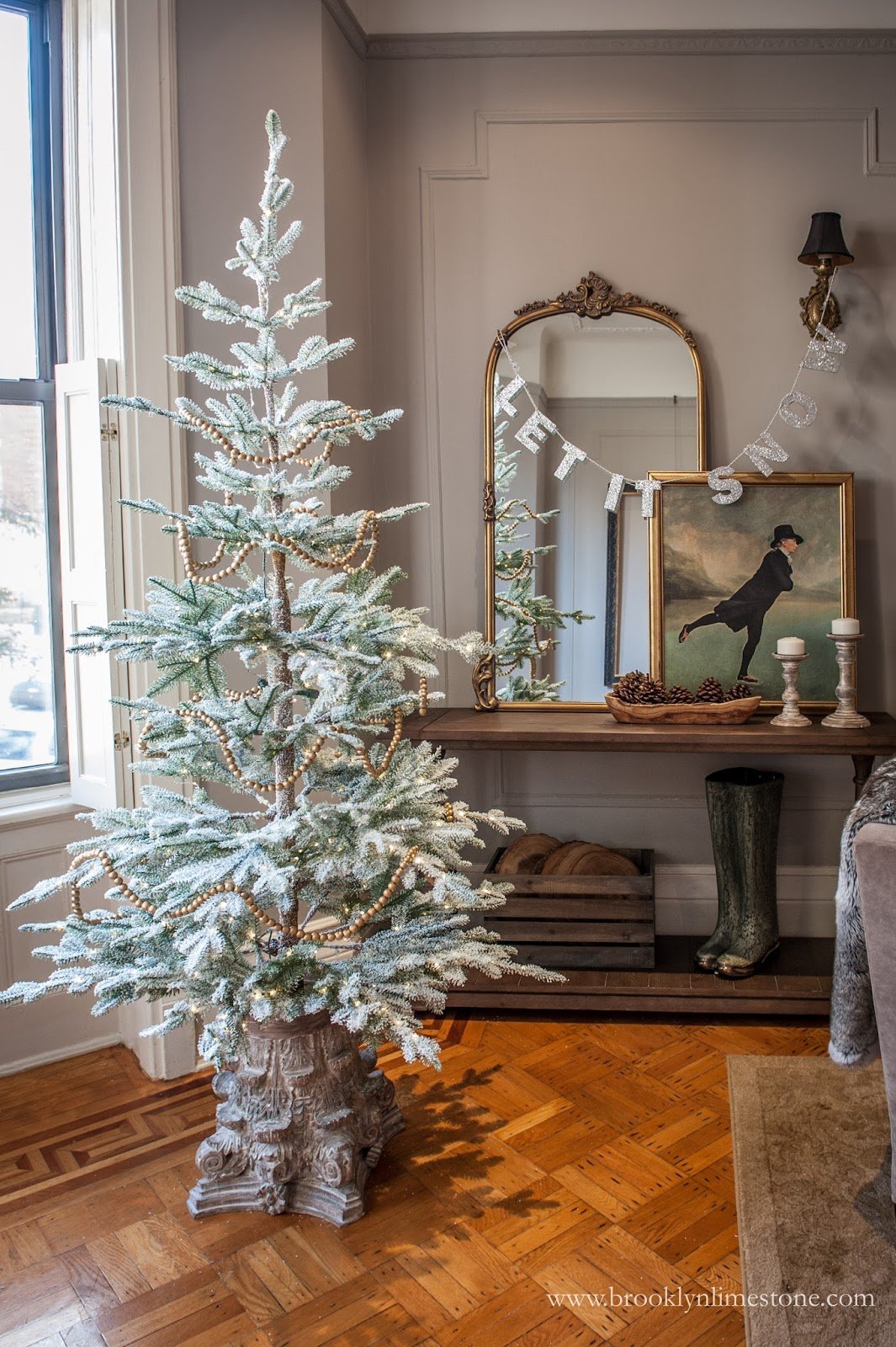 Room with a snowy artificial Christmas tree, gold beads decoration, ornate mirror with 'Let It Snow' banner, framed ice-skating painting, candles, pine cones, vintage boots, and wooden floor.