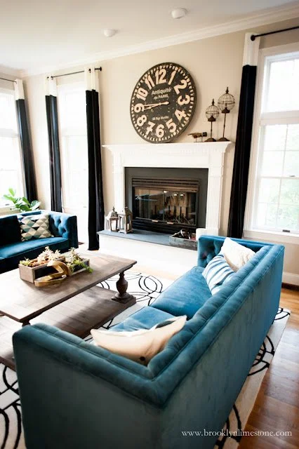 Elegant living room with blue velvet sofa set, large wall clock above white fireplace, wooden coffee table with decorative tray, and patterned rug in front of large windows with black curtains.