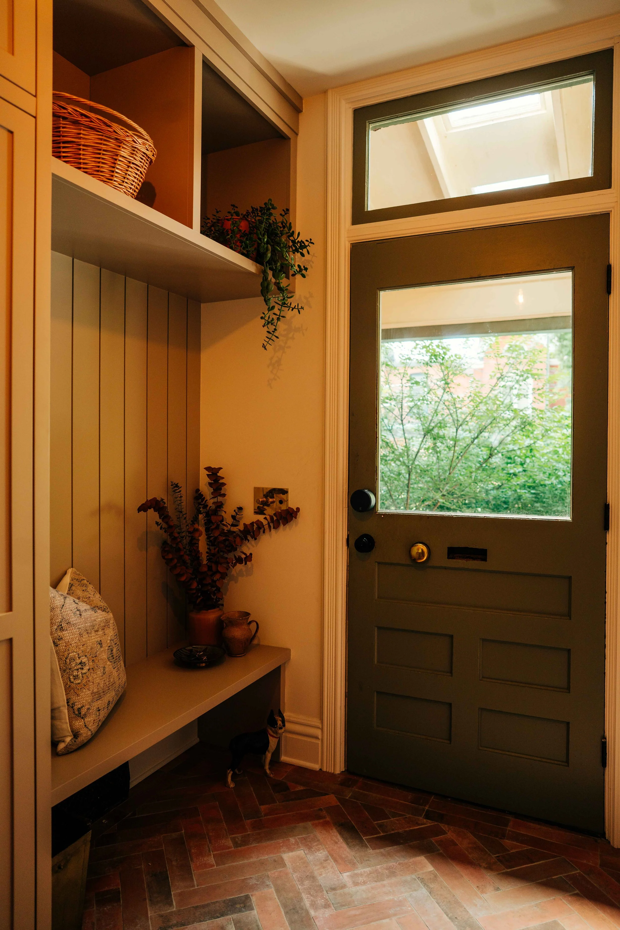 A cozy entryway with a green door featuring a window, a wooden bench with a cushion, decorative pottery, a hanging plant, and a small dog standing on the floor.