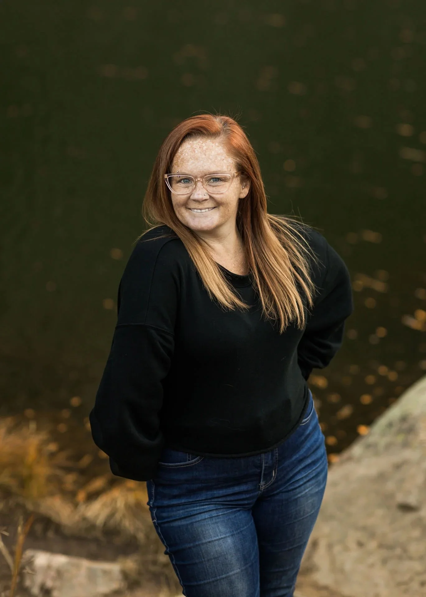 A woman with red hair, glasses, and freckles, standing outdoors near a body of water, smiling at the camera, wearing a black top and blue jeans.