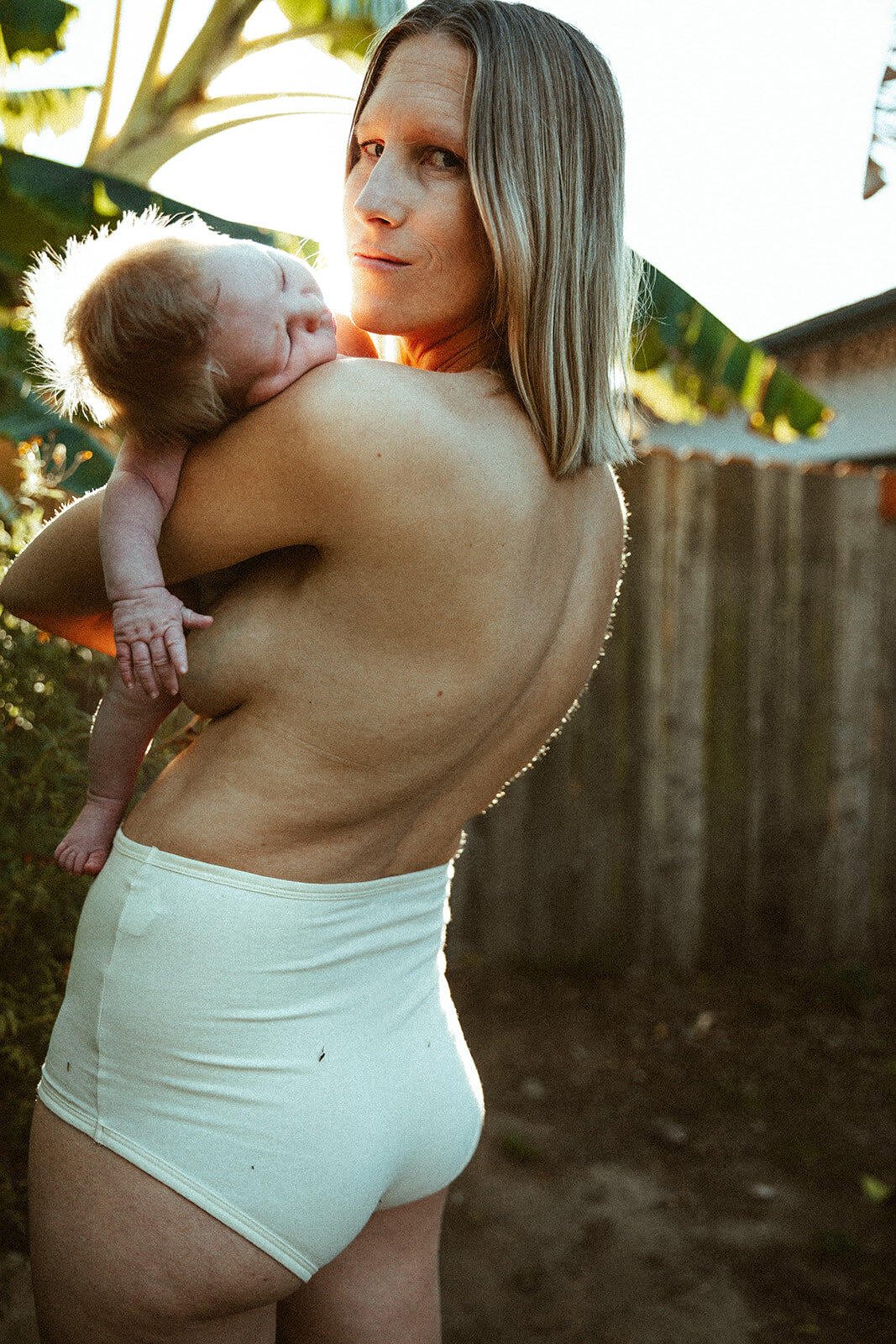 A woman holding a newborn baby outdoors during sunset, with tropical plants and a wooden fence in the background.