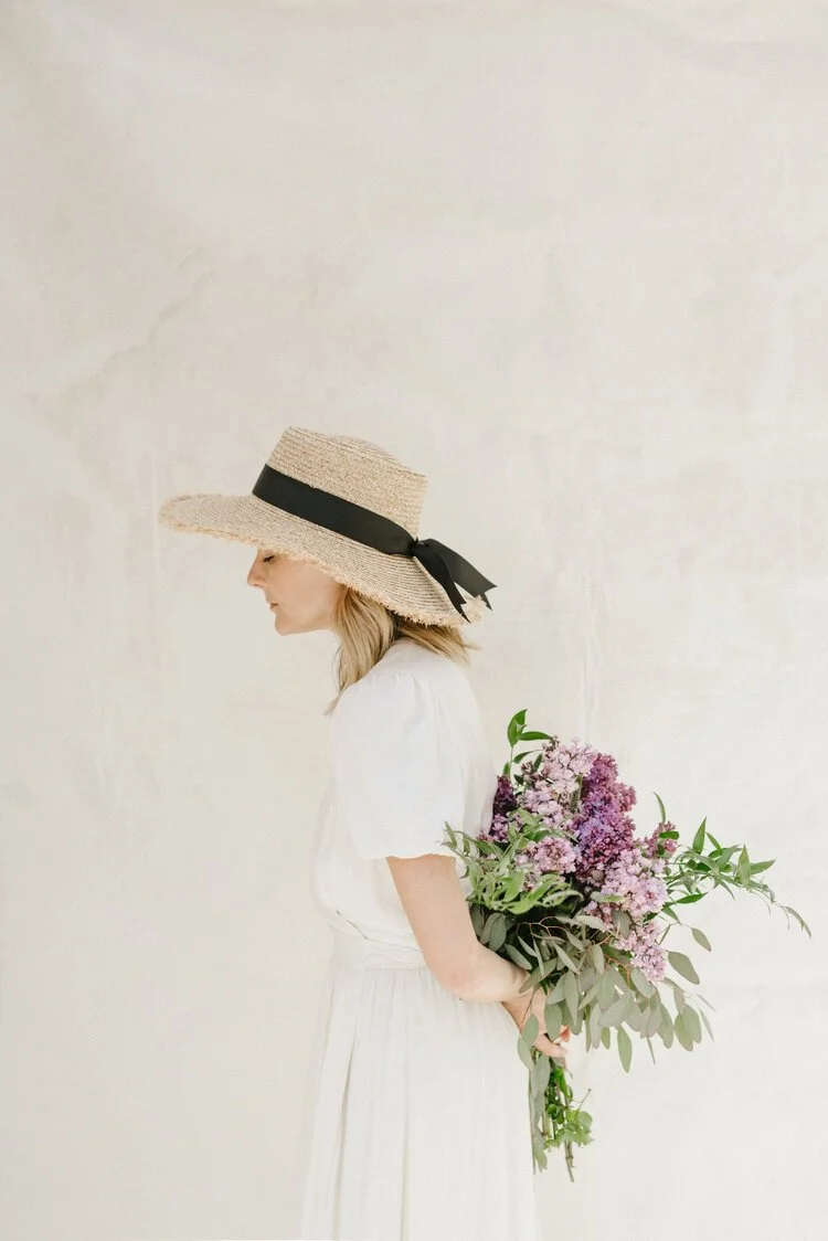 A woman wearing a wide-brimmed straw hat with a black ribbon, dressed in a white dress, is holding a bouquet of pink and purple flowers behind her back, standing against a neutral wall.