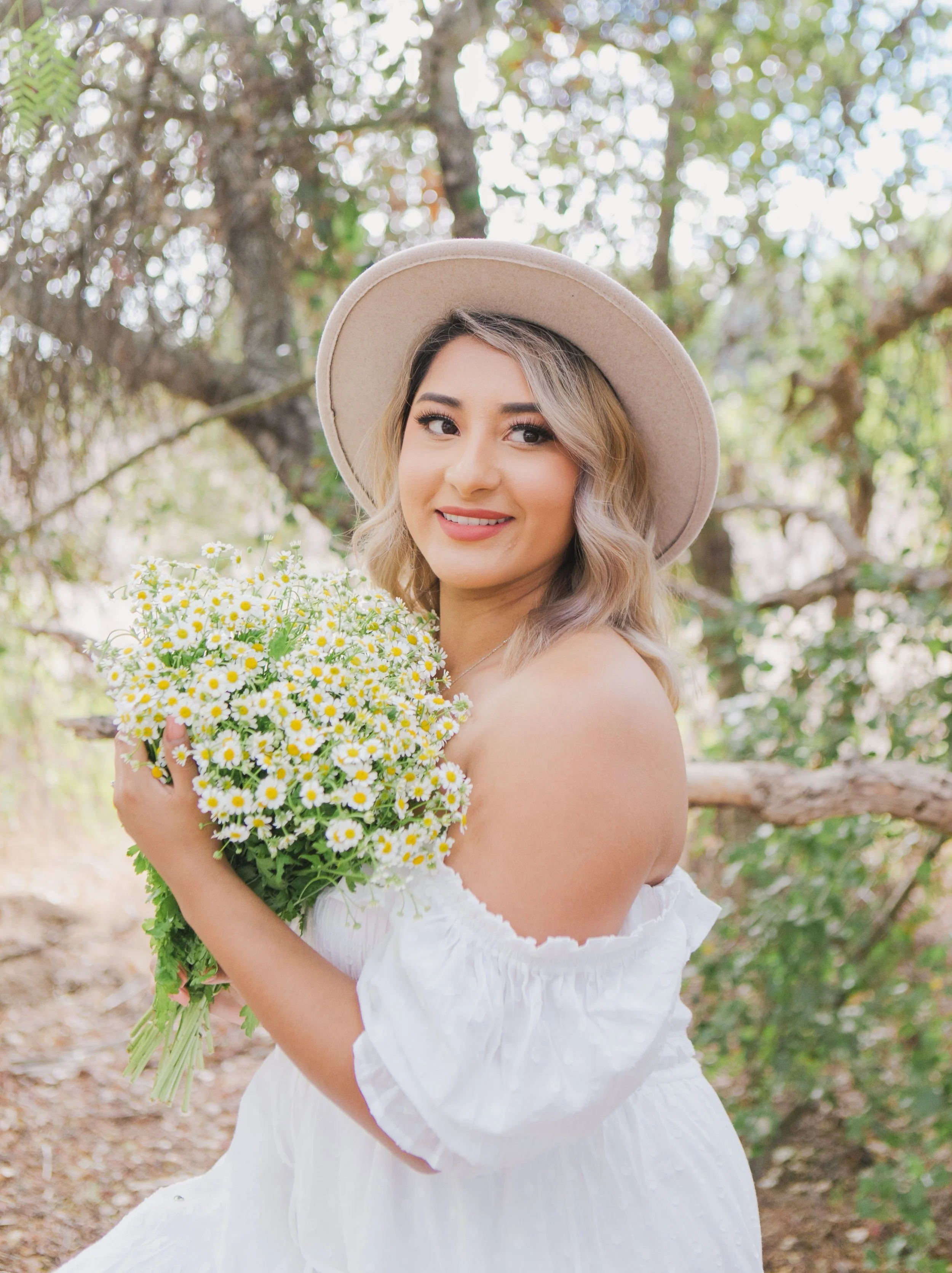A smiling woman in a white off-shoulder dress and beige hat holding a bouquet of daisies, standing outdoors among trees.