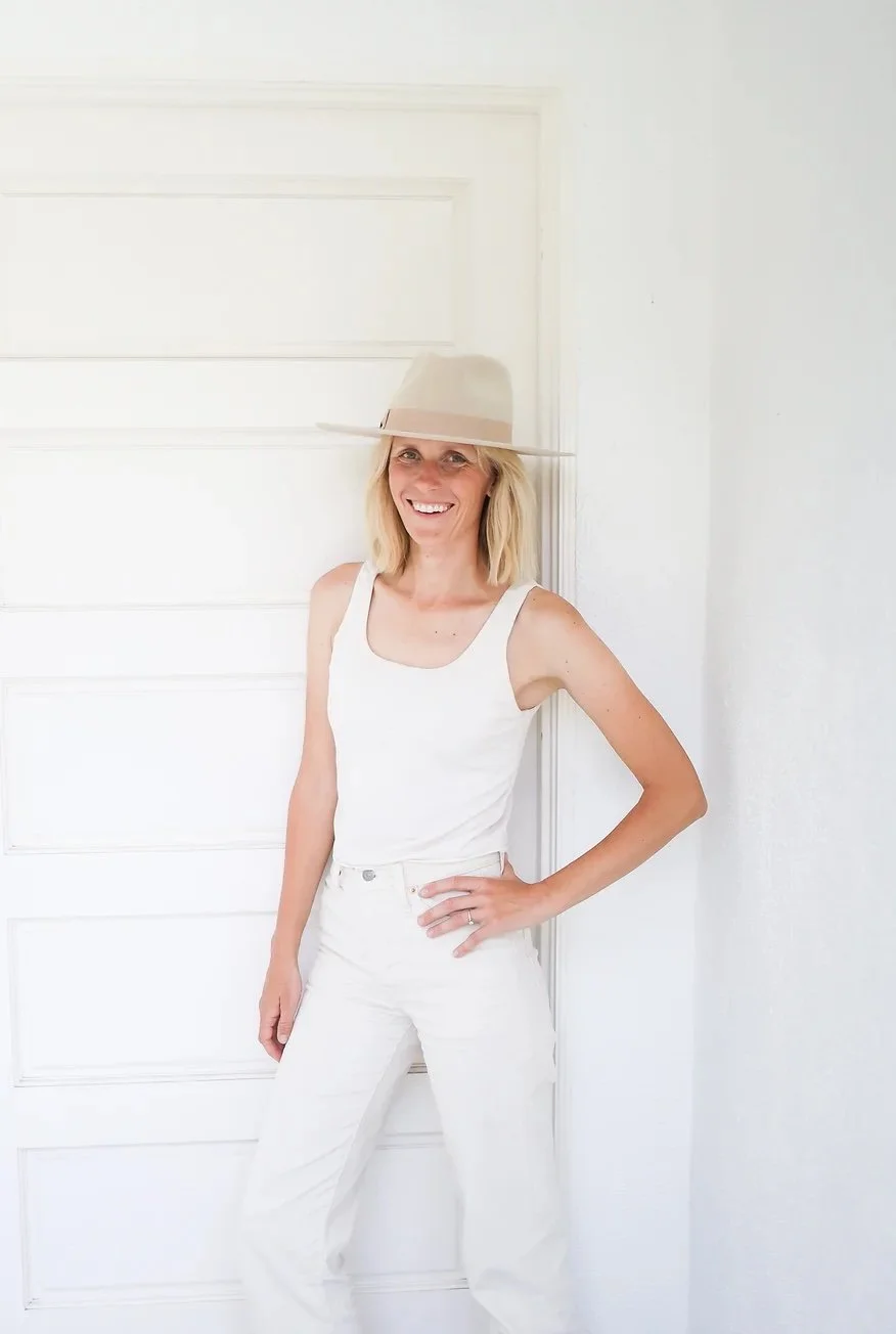 Woman in white tank top and white pants wearing a large beige hat, standing and smiling indoors against a white door and wall.