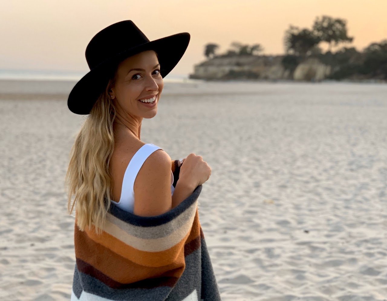 Smiling woman in a black wide-brim hat and striped blanket on a sandy beach during sunset, with cliffs and trees in the background.