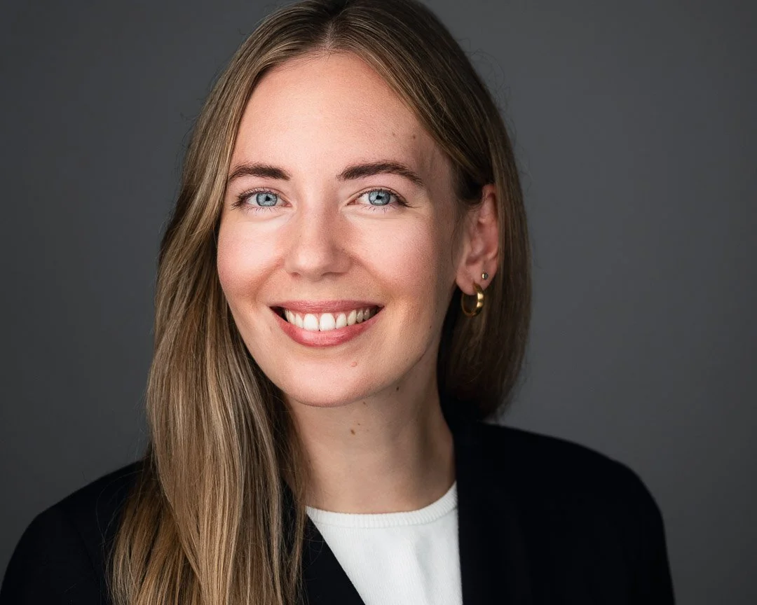 Lächelndes Bewerbungsfoto einer blonden Frau mit schwarzem Blazer vor grauem Hintergrund