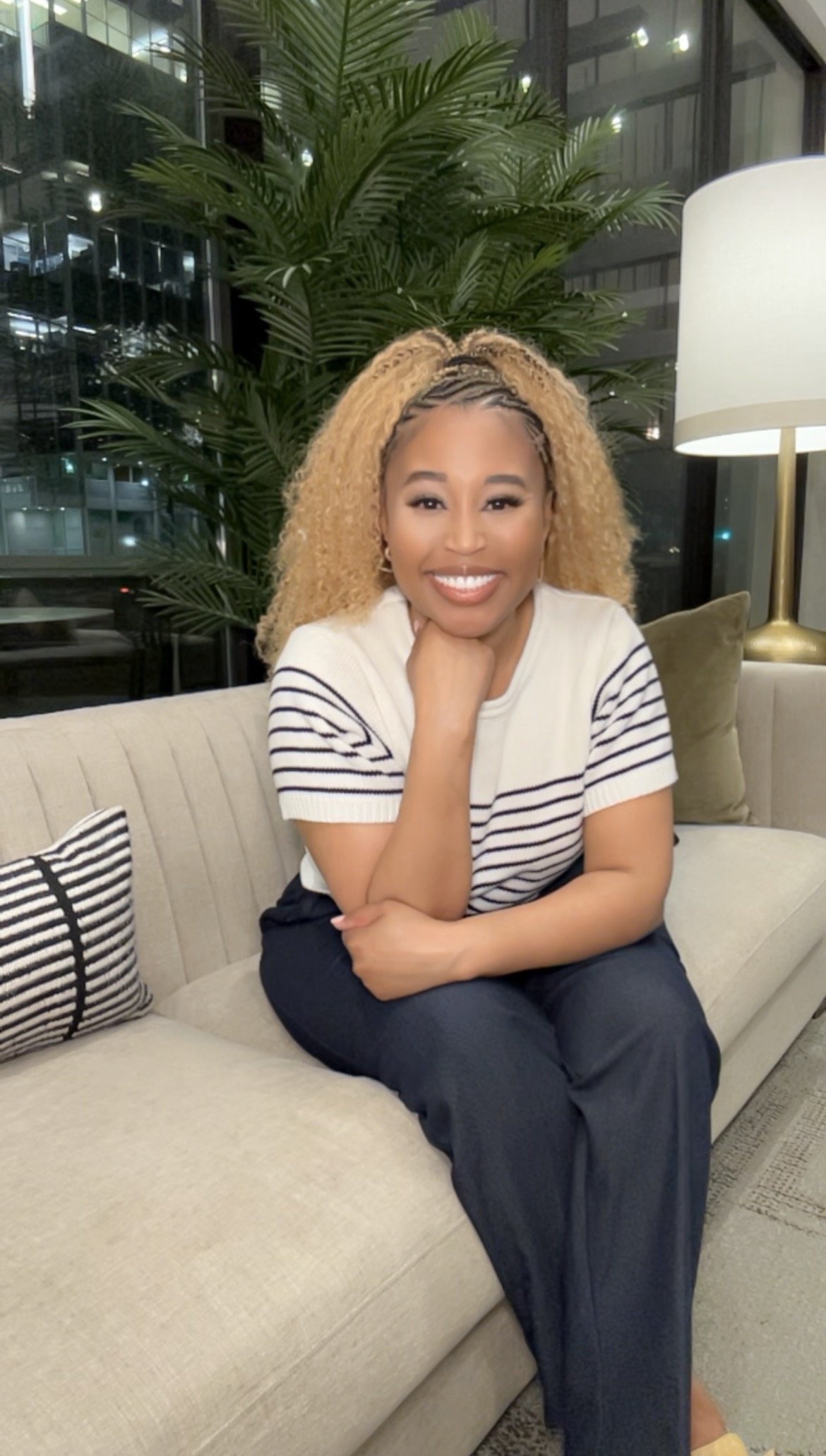 A woman with blonde curly hair, wearing a white and navy striped t-shirt and navy pants, sitting on a beige sofa in a modern indoor setting with large windows, a tall green plant, a white floor lamp with a brass base, and decorative pillows.