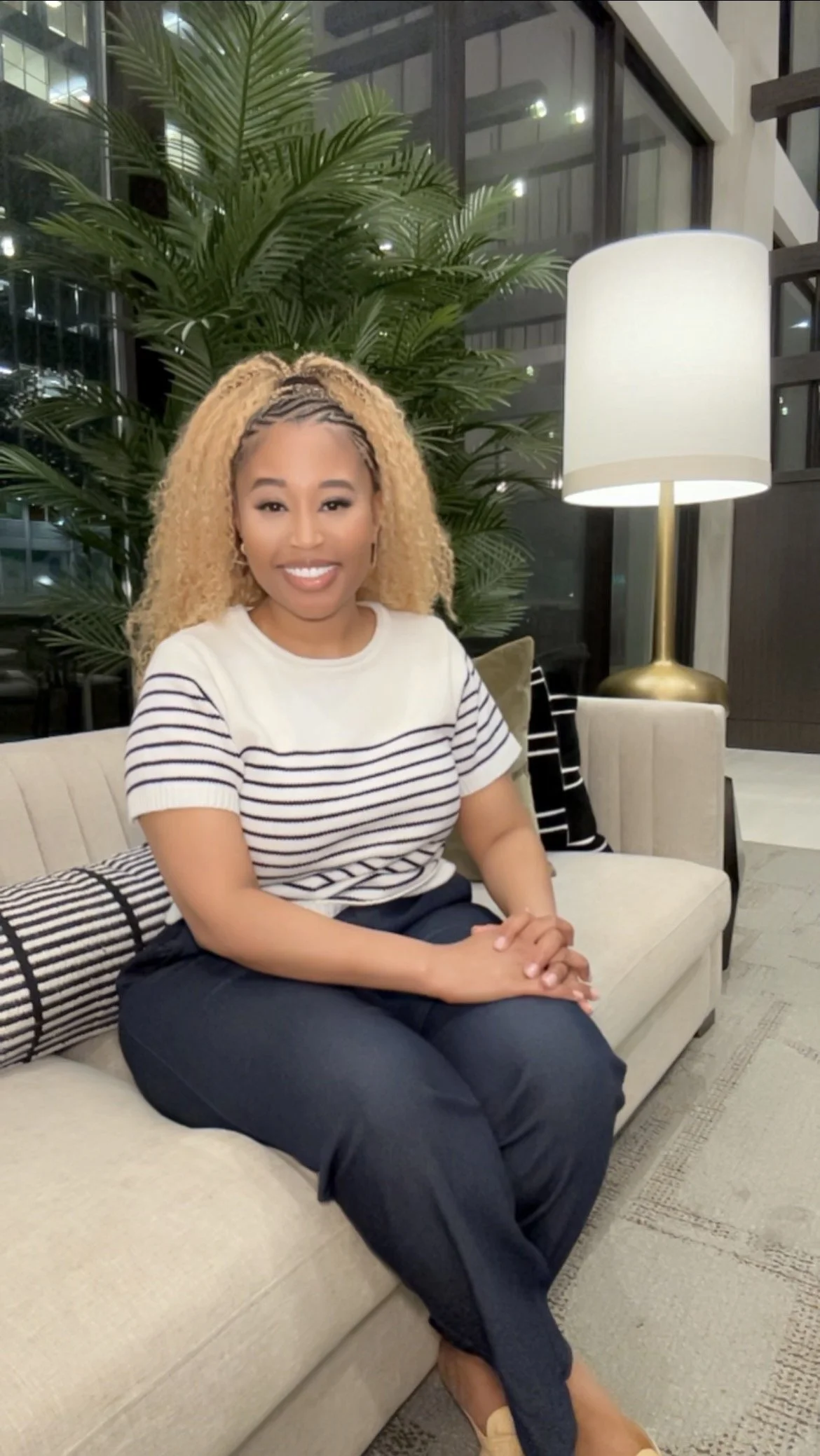 A woman with blonde, curly hair seated on a beige sofa in a lobby or lounge area. She is smiling, wearing a striped shirt and dark pants, with a large plant and a tall lamp in the background.