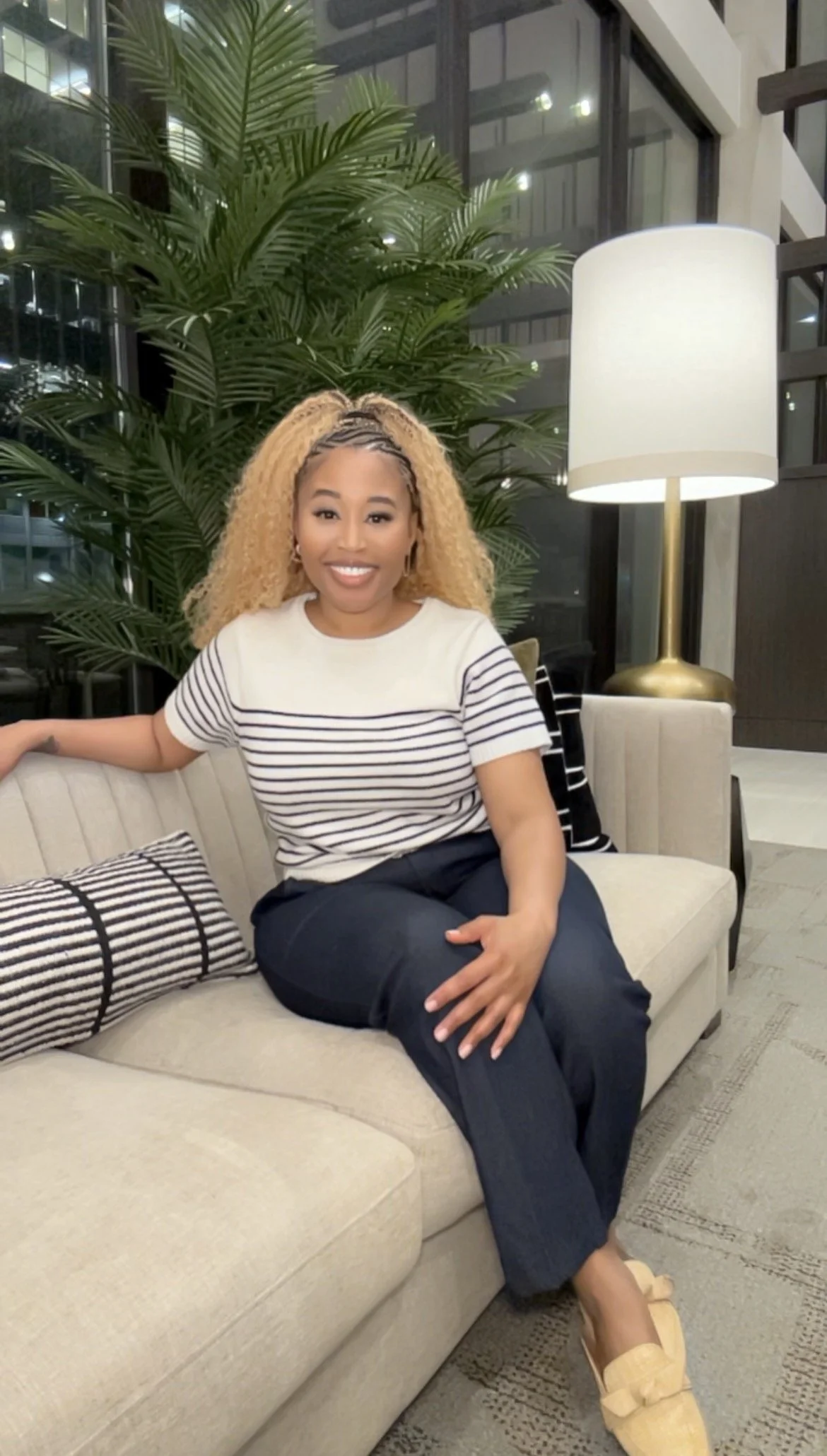 A woman with curly blonde hair, wearing a striped shirt and dark pants, sitting on a beige sofa in a modern indoor setting with large windows, a tall plant, a white lampshade, and a decorative pillow.